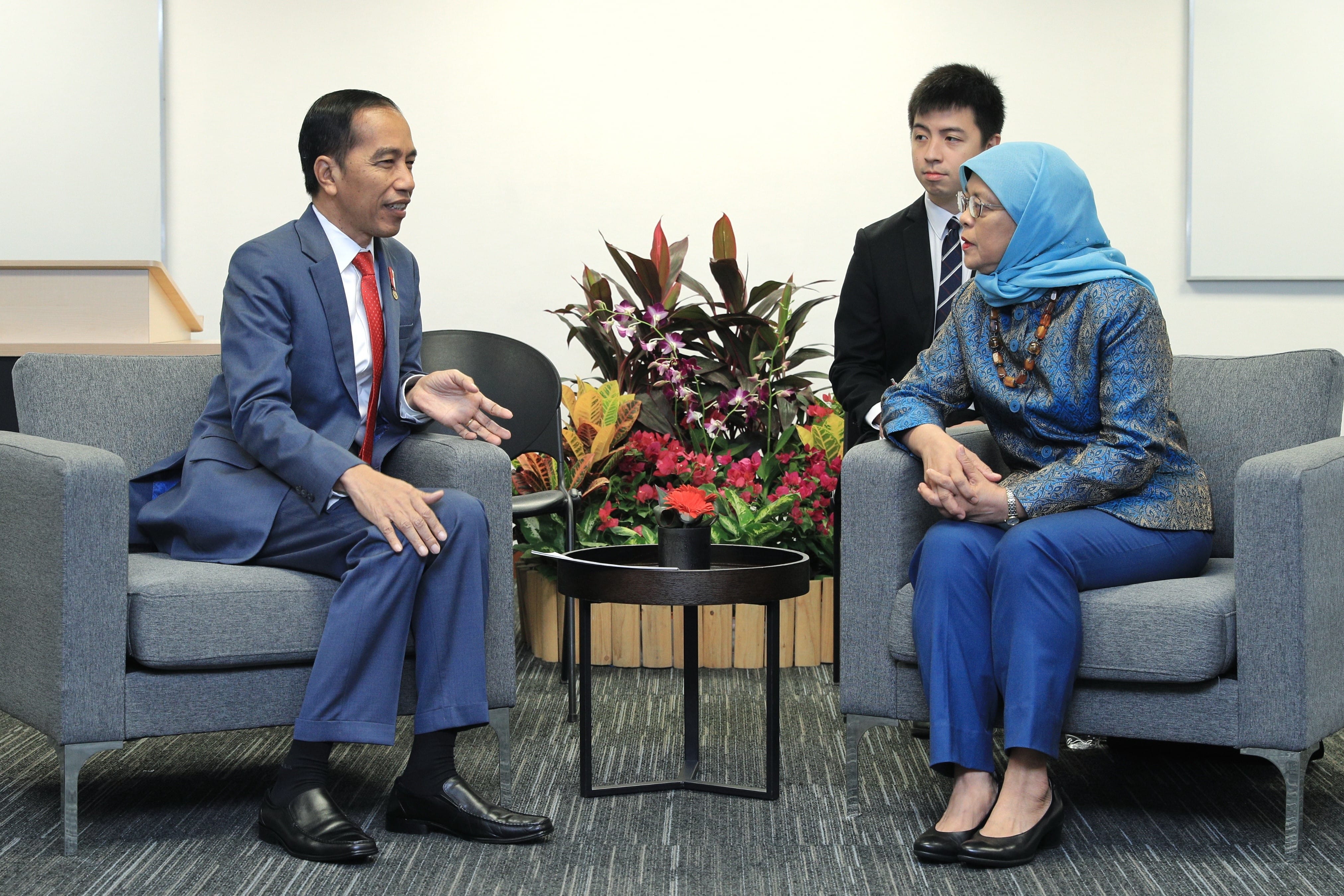 Joko Widodo with woman in blue hijab seated in gray chairs, with standing man.